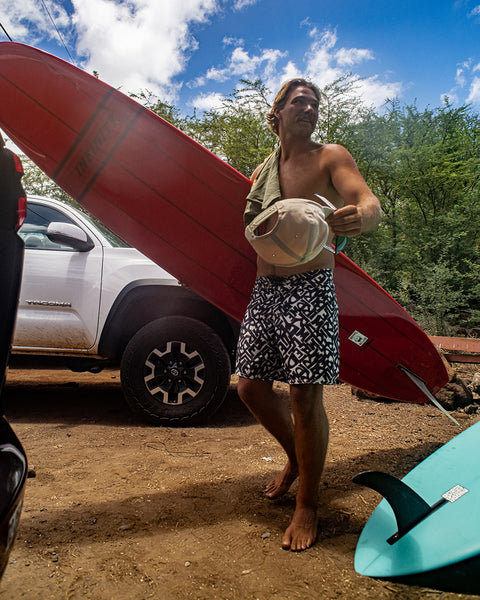 Wearing 300 Boardshorts - Alphabet Soup, a man stands barefoot on dirt by a white truck, holding a hat and towel. A red surfboard leans on the truck, with a blue surfboard in front. Trees and a partly cloudy sky are behind him.