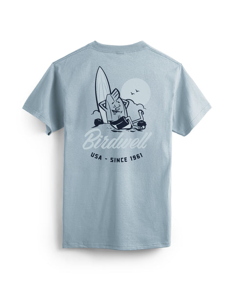 The Pau T-Shirt - Arctic Blue is a cotton heavyweight tee featuring a happy cartoon bird with a surfboard, ocean waves, and the sun. “Birdwell USA - Since 1961” text appears below the vivid illustration.