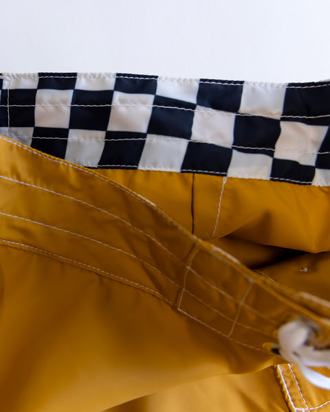 Close-up of 300 Boardshorts - Checkmate Gold: yellow SurfNyl swim trunks with white stitching, drawstring, and black-and-white checkered waistband lining. Water-repellent and made in the USA.