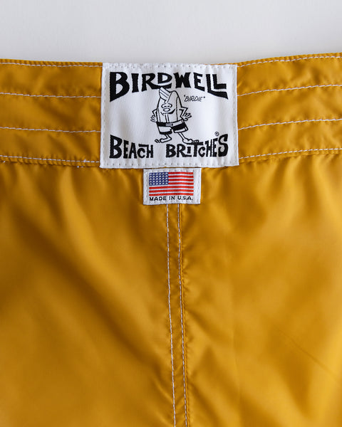 Close-up of the 300 Boardshorts - Checkmate Gold, featuring a white Birdwell Beach Britches label with a cartoon bird and a small tag below displaying the American flag and 