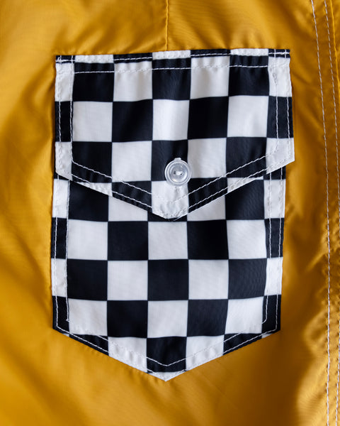 A close-up of the 300 Boardshorts - Checkmate Gold fabric, showing a pocket with a black and white checkered pattern and a silver button centered on the flap.