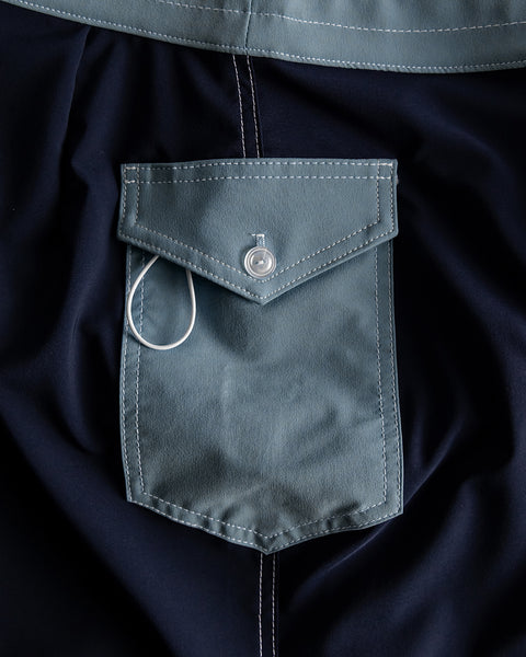 Close-up of the 808 SurfStretch Pop Pocket Boardshorts in Navy/Federal Blue, showing a flap pocket with a silver button and a white cord looped around it on dark fabric. Made in USA.