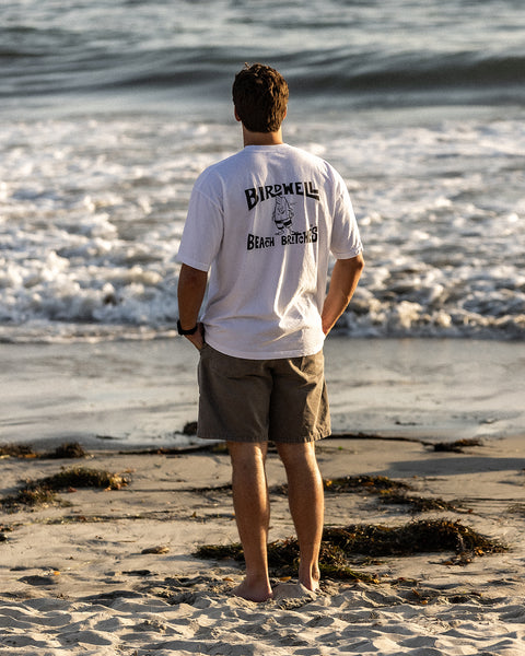 A man stands barefoot on a sandy beach, facing the ocean in gray shorts and the License Plate T-Shirt - White/Black by Birdwell Beach Britches. Waves crash behind him with seaweed scattered across the sand.