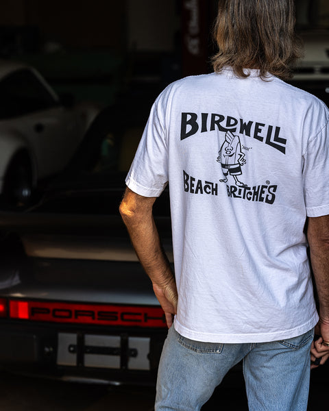 A person with long hair wearing a License Plate T-Shirt - White/Black and jeans stands in front of a black Porsche, facing away from the camera in a garage setting.
