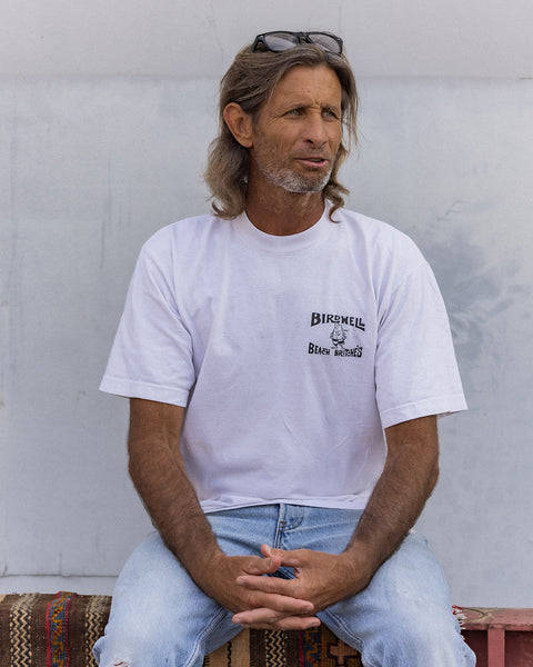 A middle-aged man with long hair and sunglasses on his head sits on a bench, wearing the License Plate T-Shirt - White/Black in heavyweight cotton and light blue jeans. He looks slightly to the side, hands clasped in his lap.