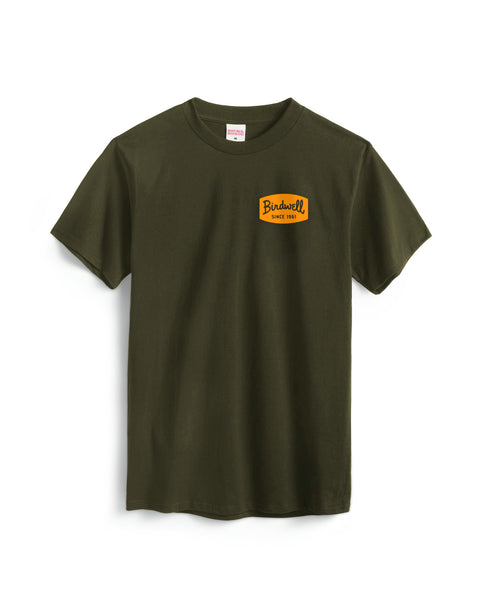 The Herald T-Shirt - Surplus Green is a heavyweight, garment-dyed dark green cotton tee with a small yellow rectangle patch reading “Bindwell Since 1983” in black on the left chest, shown against a white background.