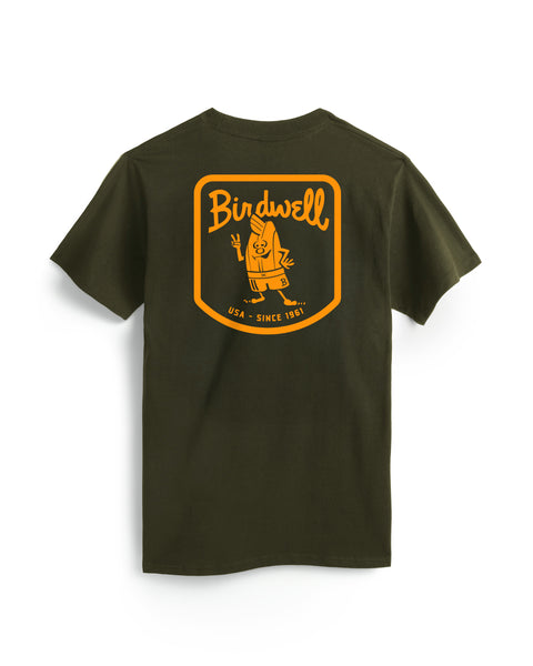 The Herald T-Shirt - Surplus Green is a dark green, garment-dyed heavyweight cotton tee with a large yellow-orange back graphic featuring “Birdwell,” a cartoon character, and “USA • SINCE 1961” in a rounded rectangle.