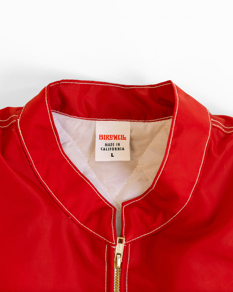 A close-up of the Quilt Lined Racing Jacket - Red/White/Black shows white stitching, a gold zipper, and a collar label that reads BIRDWELL MADE IN CALIFORNIA L.