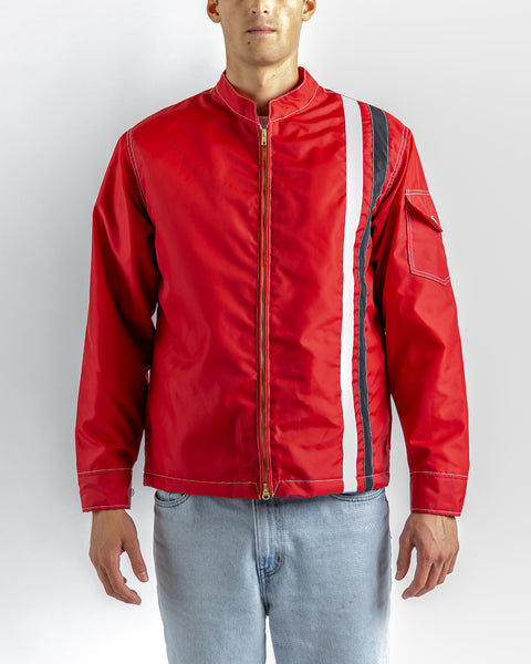 A person wearing the Quilt Lined Racing Jacket - Red/White/Black, featuring a front zipper and vertical white and black stripes on the left side, pairs it with light blue jeans while standing against a plain light gray background.