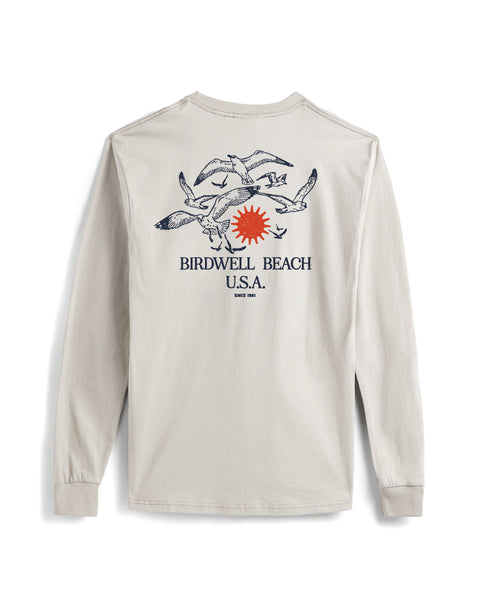 The Tourist Long Sleeve Pocket T-Shirt - Cement is a heavyweight, light beige cotton tee with seagulls, a red sun, and blue “Birdwell Beach U.S.A. Since 1961” text on the back. Features a pocket and is proudly made in the USA.