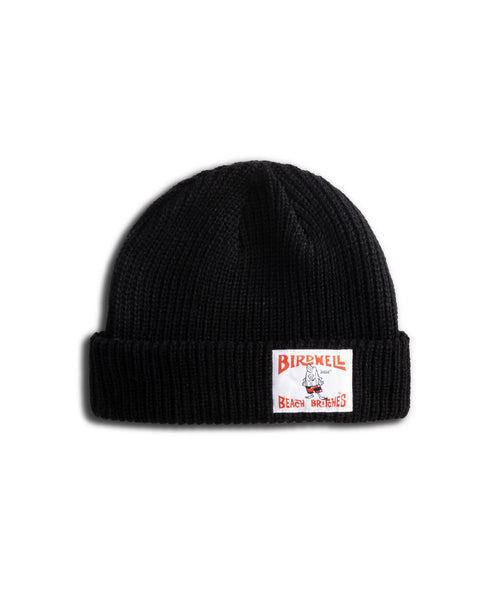 The License Plate Watchcap - Black is a classic fisherman-style beanie in black wide rib with a folded brim, featuring a rectangular white patch with red Birdwell Beach Britches text and an illustrated bird logo on the front.