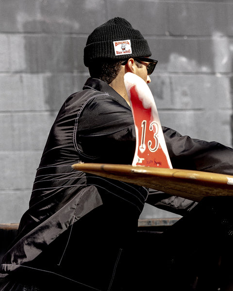 A person in a black License Plate Watchcap and sunglasses sits at an outdoor table; a red and white table marker with the number 13 is in front, with a gray brick wall in the background.