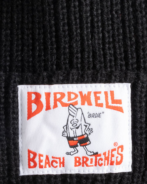 Close-up of the License Plate Watchcap - Black, a wide ribbed beanie featuring a Birdwell patch label with a cartoon character in red shorts, “BIRDWELL BEACH BRITCHES” in bold red letters, and “BIRDIE” above the character.