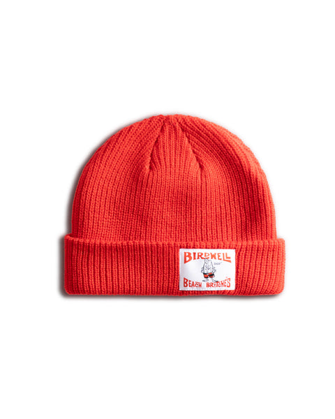 The License Plate Watchcap - Red is a bright red ribbed beanie with a folded brim, featuring a white rectangular Birdwell Beach Britches patch and an illustrated cartoon character on the front.