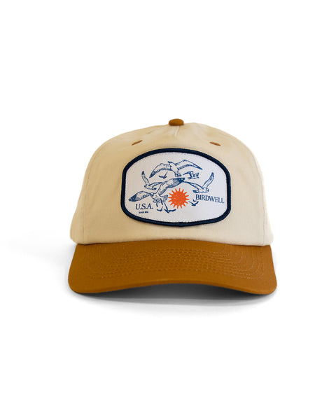 The Tourist Twill Snapback - Bone/Brown is a beige and brown cotton hat with a brown brim, front sewn patch displaying blue fish, 