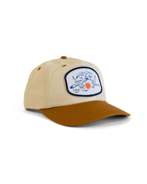 The Tourist Twill Snapback - Bone/Brown is a beige 5-panel cotton hat with a brown brim and a sewn patch showing blue hands shaking, a red sun, and the words U.S.A. and SEVISUAL. Set against a white background.