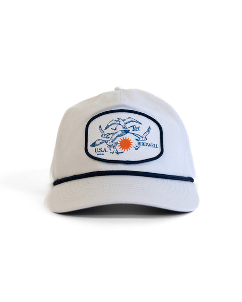 The Tourist Twill Snapback - White is a classic cotton 5-panel snapback hat with navy trim, featuring an embroidered flying fish, orange sunburst, and “U.S.A. Birdwell” text on the front.