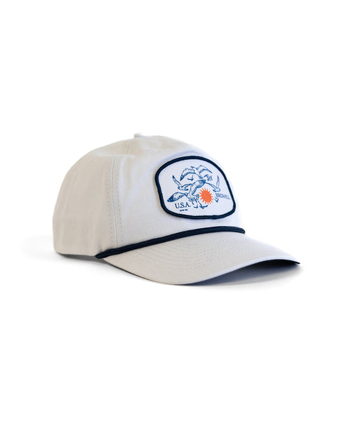 The Tourist Twill Snapback - White is a light-colored 5 panel hat with a blue crab and red sun patch on the front, plus dark piping along the brim.