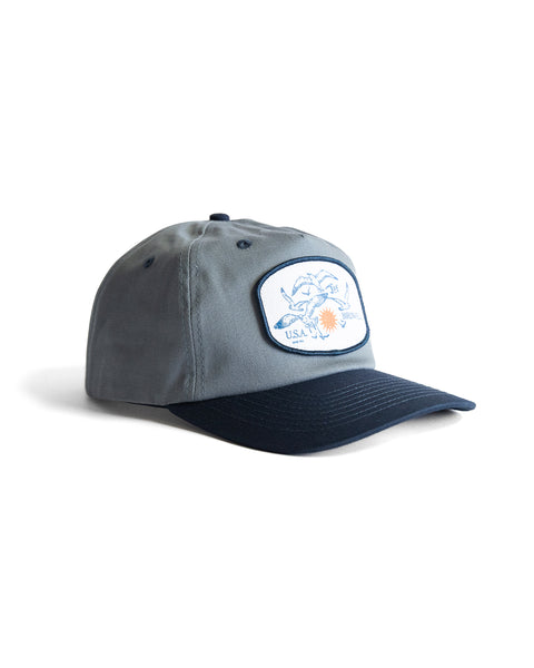 The Tourist Twill Snapback - Federal Blue/Navy is a 5-panel gray and navy blue cotton twill hat with a curved brim, ventilation eyelets, and a front patch showing a blue crab and the text USA and Sunshine.