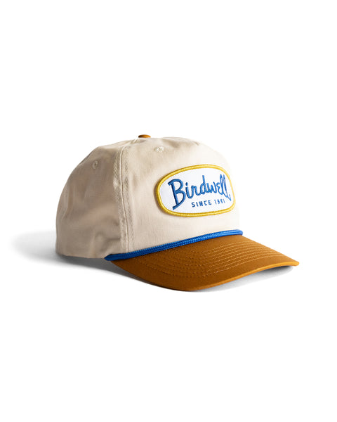 The Potato Patch Twill Snapback - Bone/Brown is a beige 5-panel hat with a brown brim and blue trim, featuring an oval 