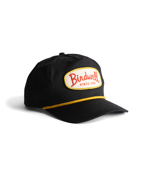The Potato Patch Twill Snapback - Black is a 5-panel unstructured snapback hat with a curved brim, yellow trim, and a white front patch displaying 