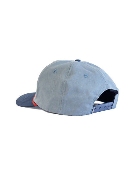 The Milkman Twill Snapback in Federal Blue/Navy is a medium crown cotton 5-panel hat with a navy brim and red accents. It has an adjustable snapback closure, ventilation eyelets, and is pictured from the back on a white background.