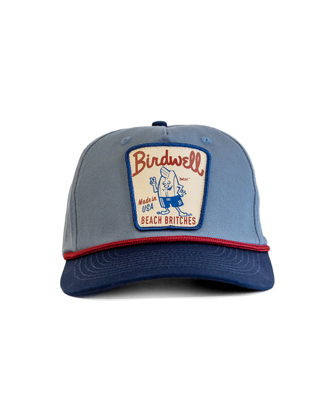 The Milkman Twill Snapback - Federal Blue/Navy is a classic 5-panel cotton hat with a Birdwell front patch, cartoon figure, “Made in USA” and “Beach Britches” text, plus red trim accenting the brim and crown.