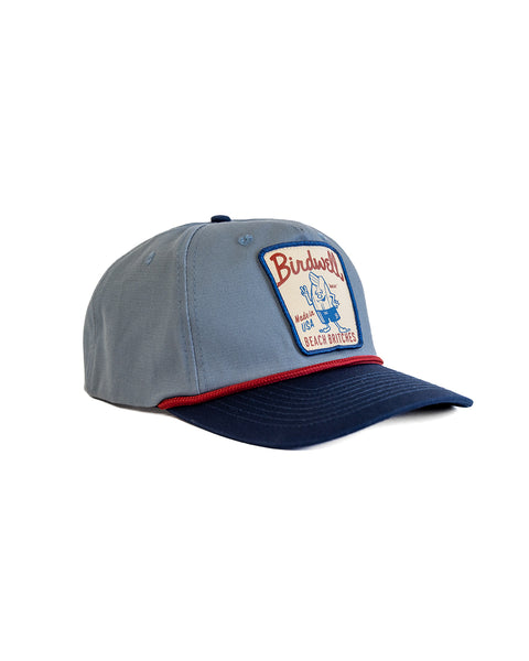 The Milkman Twill Snapback in Federal Blue/Navy is a cotton 5-panel hat with a navy brim, red trim, and medium crown fit. The front patch says Birdwell Beach Britches with a swimwear character holding a flag.