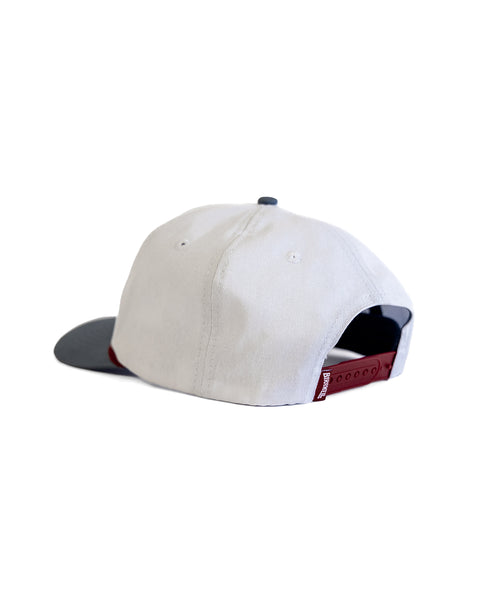 The Milkman Twill Snapback - White/Federal Blue is a white cotton 5-panel hat featuring a dark gray brim and red adjustable snap closure, displayed from the back against a white background.