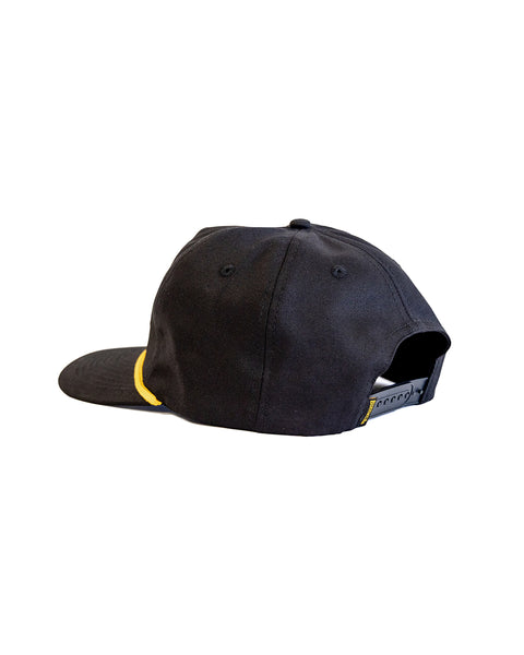 The Shield Twill Snapback - Black is a black cotton high crown hat with a yellow side accent, adjustable strap, and ventilation eyelets, shown from the back against a white background.