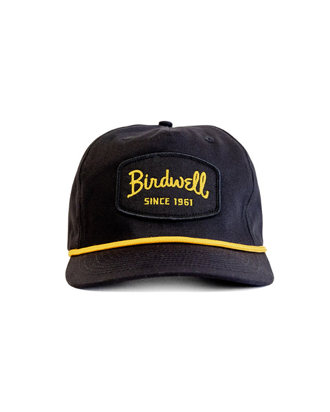 The Shield Twill Snapback - Black is a cotton hat with a yellow embroidered 