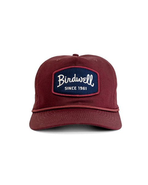 The Shield Twill Snapback - Burgundy is a 5-panel maroon cotton hat with a navy front patch reading 