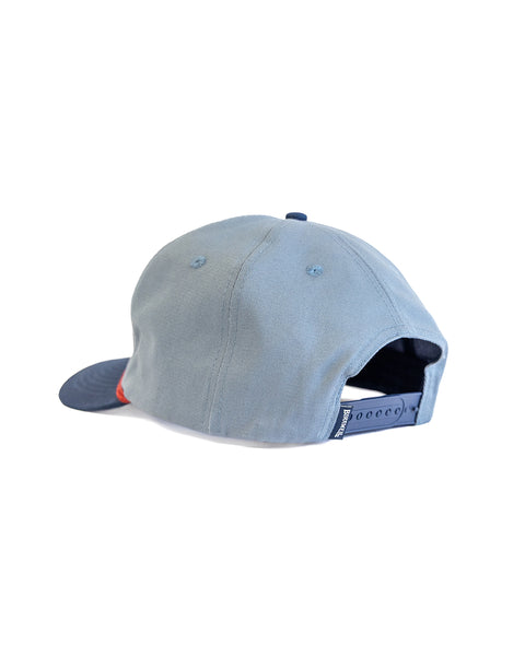 The Shield Twill Snapback - Federal Blue/Navy is a light blue 5-panel hat shown from the back, featuring a navy adjustable strap and small ventilation holes on top, set against a white background.