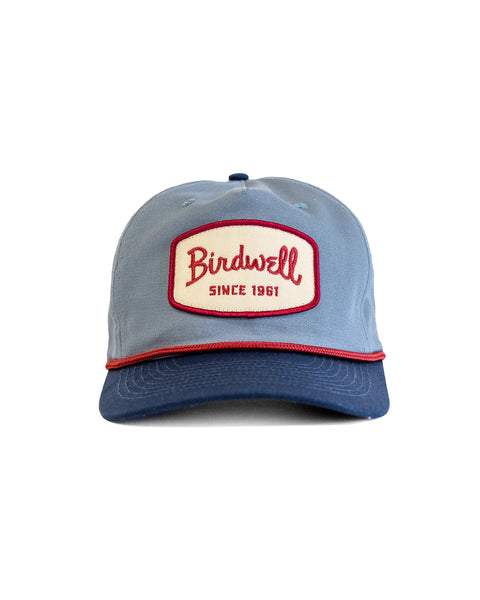The Shield Twill Snapback - Federal Blue/Navy is a light blue cotton hat with a navy brim, red piping, and a white patch with red trim displaying 