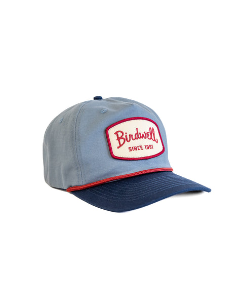 The Shield Twill Snapback in Federal Blue/Navy is a 5-panel hat with a navy brim and a red-trimmed front patch reading 