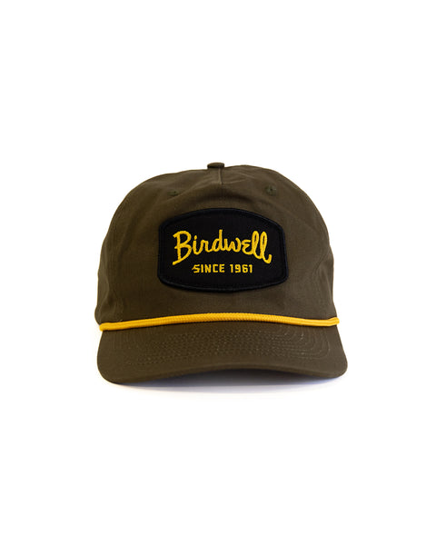 The Shield Twill Snapback - Olive is a high crown, 5 panel cotton hat in olive green with a black and yellow 
