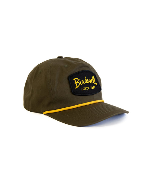 The Shield Twill Snapback - Olive is a 5-panel cotton snapback hat in olive green with a curved brim, yellow accent line, and a high crown front featuring a black patch with yellow 