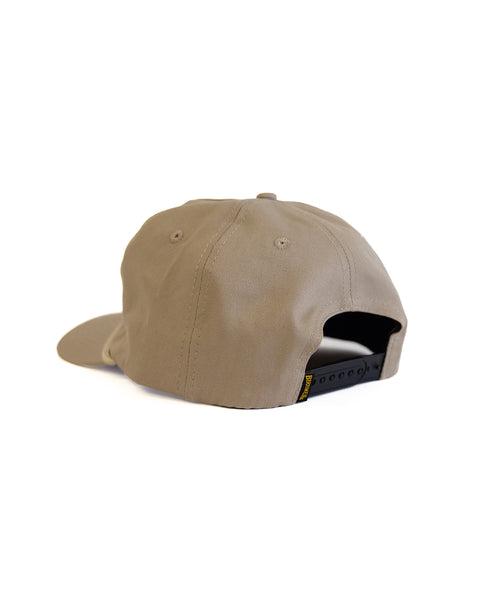 The Shield Twill Snapback - Tan is shown from the back, featuring a black adjustable strap and stitched panels, set against a plain white background.