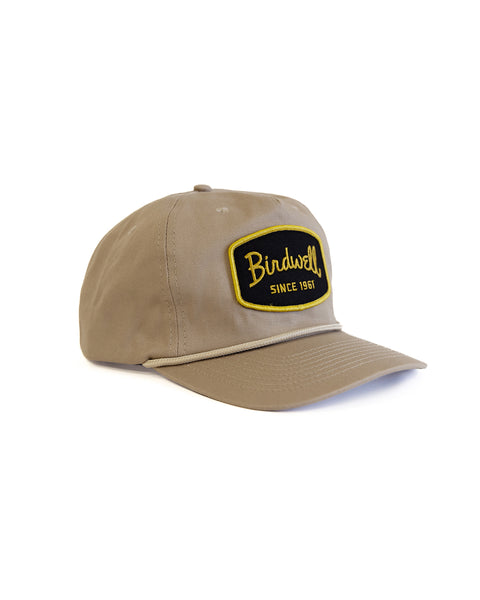 The Shield Twill Snapback - Tan is a khaki cotton hat with a curved brim and a black and yellow embroidered patch reading 