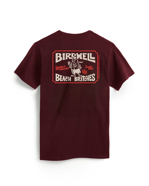 The Hangtag T-Shirt - Burgundy is a heavyweight, garment-dyed cotton tee with short sleeves and a bold vintage-style graphic on the back showing Birdwell Beach Britches and a surfboard-carrying figure.
