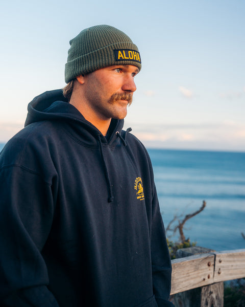 A mustachioed man outdoors by a wooden fence, with ocean and sky behind him, wears a black hoodie and the Aloha Watchcap - Army, a green ribbed fisherman beanie featuring an Aloha patch.