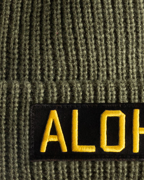 Close-up of the Aloha Watchcap - Army, a green ribbed beanie with a black patch displaying 