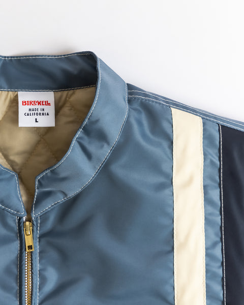 Close-up of the Quilt Lined Racing Jacket in Federal Blue/Bone/Navy with cream and navy stripes, gold zipper, and a white BIRDWELL MADE IN CALIFORNIA L label inside the collar.