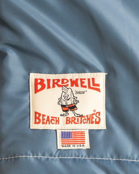 A label on the Quilt Lined Racing Jacket - Federal Blue/Bone/Navy features Birdwell Beach Britches, a cartoon character, and a small American flag patch below, proudly stating 