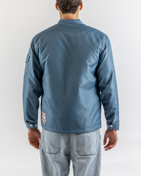 A man faces away from the camera, wearing the Quilt Lined Racing Jacket in Federal Blue/Bone/Navy with a patch on the lower back, sleeve pocket, and light blue jeans. The scene is set against a plain, light-colored background.