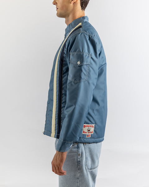 A man stands in profile wearing the Quilt Lined Racing Jacket in Federal Blue/Bone/Navy, featuring a white vertical stripe and sleeve patch pocket, paired with light blue jeans against a plain background.