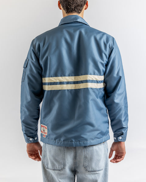 A person faces away, wearing the Comp Jacket 25 in Federal Blue/Bone with two beige stripes across the back and light blue jeans. A text patch with a small flag is near the hem, highlighting classic SurfNyl durability against a white background.