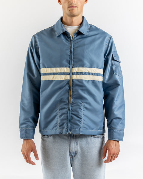 A man wears the American-made Comp Jacket 25 in Federal Blue/Bone, featuring two white horizontal stripes, paired with light blue jeans, standing against a plain white background.