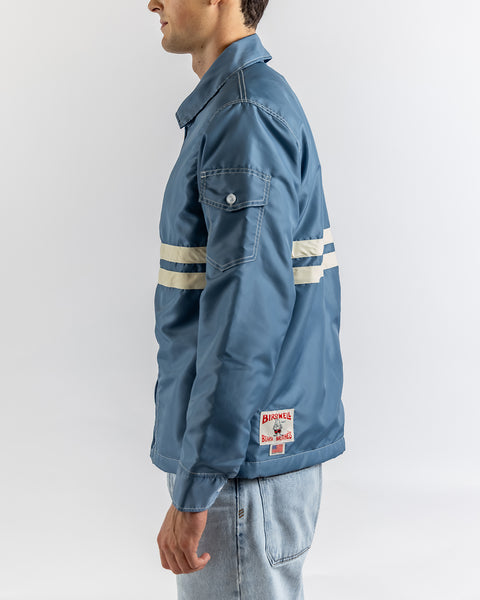 A person wears the Comp Jacket 25 in Federal Blue/Bone, featuring white horizontal sleeve and back stripes, SurfNyl durability, and a Bushnell Blue Sands patch at the hem. It's styled with light blue jeans.
