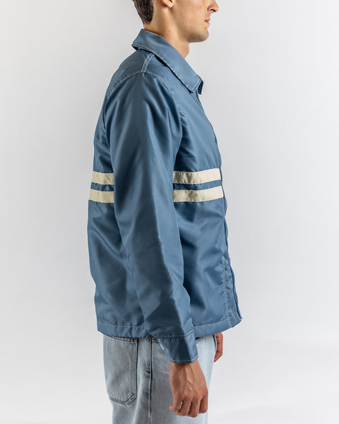 Wearing the Comp Jacket 25 - Federal Blue/Bone—crafted in the USA and known for SurfNyl durability—someone in light blue jeans stands sideways, highlighting its striking blue color and bold white stripes on a plain white backdrop.